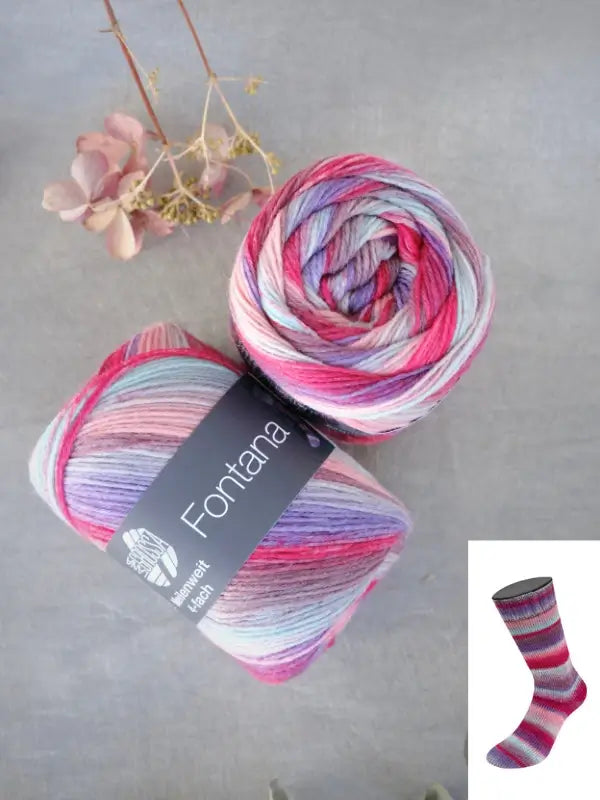 Two rolls of Lana Grossa Meilenweit 100 Fontana yarn in pastel multicolors—including pink, purple, and blue—are pictured, with a dried flower placed above them. A small inset displays a knitted sock made from this yarn.
