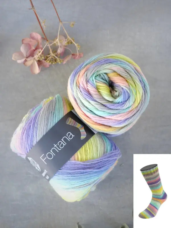 Two skeins of Lana Grossa Meilenweit 100 Fontana yarn in pastel rainbow hues are arranged on a gray surface beside dried flowers. An inset features a sock knitted from this yarn, showcasing its vibrant striped pattern.