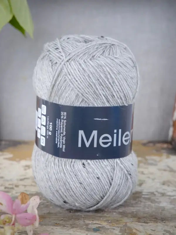 A ball of Lana Grossa Meilenweit 100 Tweed yarn in light gray is positioned upright on a worn, textured surface, accompanied by a small pink flower in the corner and a green leaf in the background, highlighting the premium quality characteristic of this yarn.