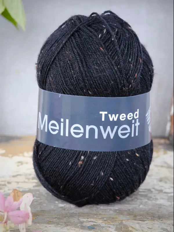 A skein of Lana Grossa Meilenweit 100 Tweed yarn in black, featuring small colored flecks, rests on a distressed wooden surface.