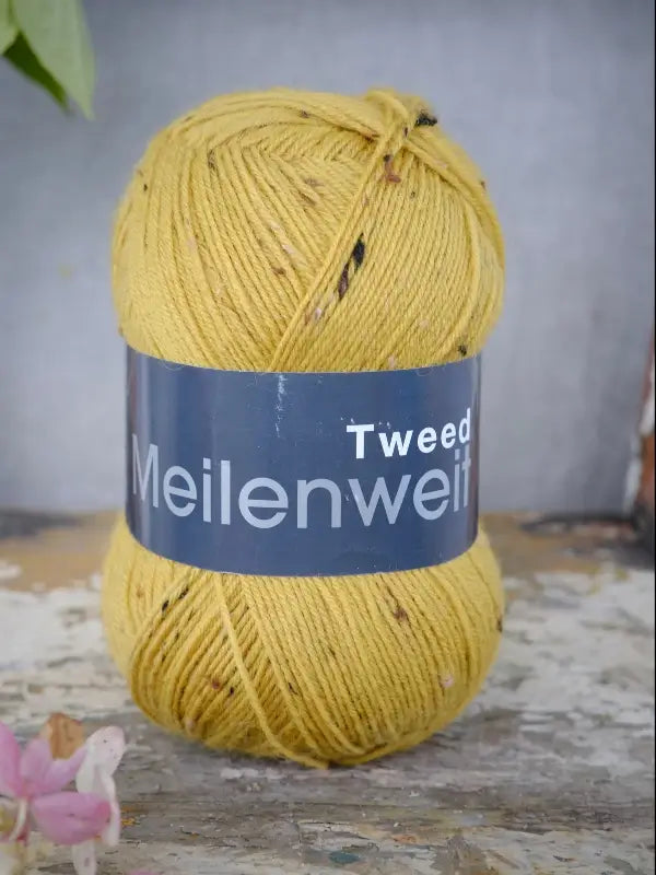 A skein of Lana Grossa Meilenweit 100 Tweed yarn in yellow rests on a worn wooden surface, with a blurred background and a small pink flower in the corner.