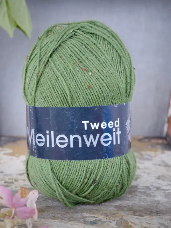 A skein of Lana Grossa Meilenweit 100 Tweed green yarn rests on a rustic surface, with a blurred background and a small pink flower in the foreground—perfect for your next sukkalanka project.
