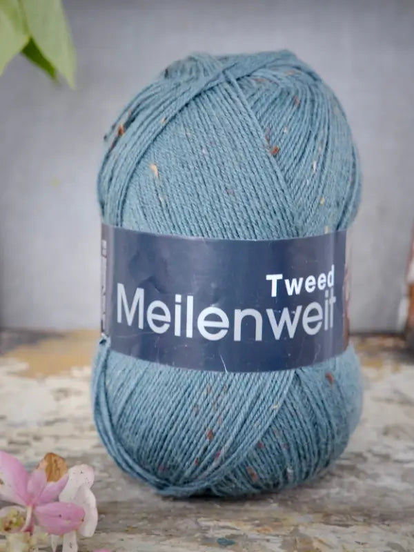 A skein of Lana Grossa Meilenweit 100 Tweed yarn in blue, featuring subtle multicolored flecks, rests on a rustic surface. A small pink flower in the lower left corner adds a delicate touch to the sukkalanka display.