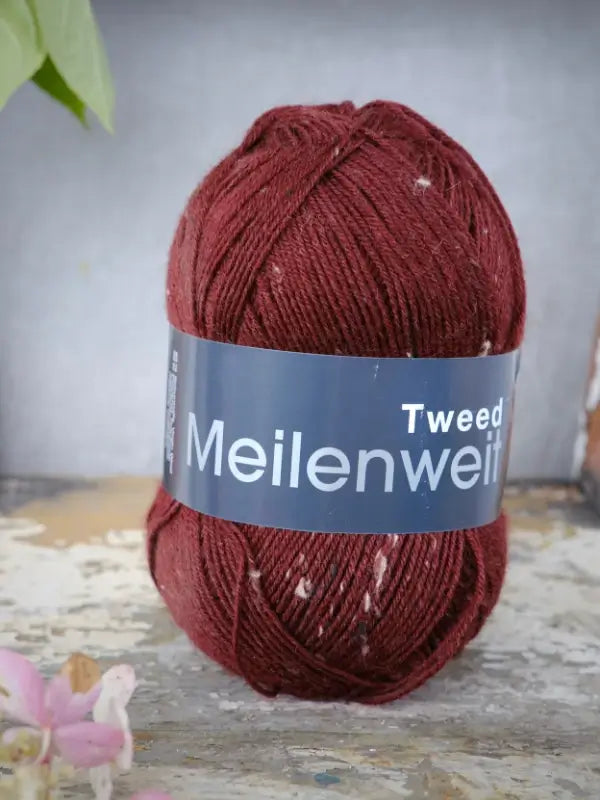A skein of Lana Grossa Meilenweit 100 Tweed sock yarn in red with white flecks rests on a rustic wooden surface, with a blurred plant and pink flower in the background.