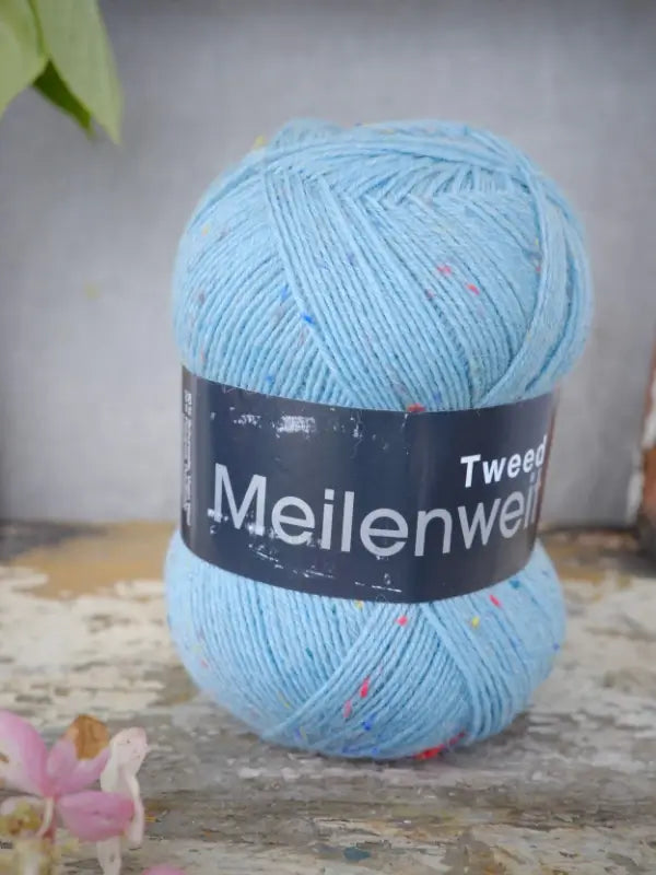 A ball of Lana Grossa Meilenweit 100 Tweed yarn in light blue, flecked with tiny colored specks, sits atop a rustic, worn wooden surface. Soft pink flowers peek into the lower left corner, bringing a gentle touch to the scene. This premium sukkalanka is ideal for your next cozy project.
