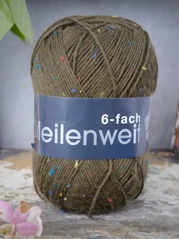 A skein of Lana Grossa Meilenweit 6 Tweed yarn in olive green with colorful flecks is displayed on a rustic wooden surface, accompanied by a small pink flower in the corner, emphasizing the charming appearance of this kuusisäikeinen tweed-sukkalanka.