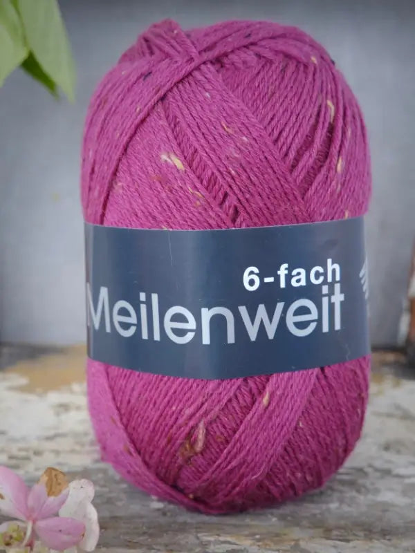 A skein of Lana Grossa Meilenweit 6 Tweed in magenta, featuring delicate light flecks, stands upright on a rustic surface with a small pink flower placed nearby.