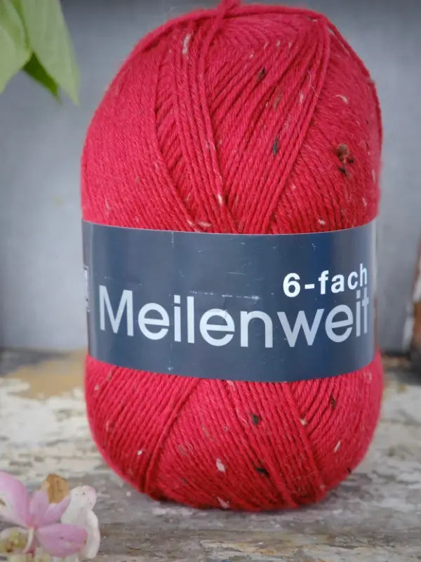 A skein of Lana Grossa Meilenweit 6 Tweed red yarn rests on a worn surface, accompanied by a small pink flower, highlighting the beautiful texture of this kuusisäikeinen sukkalanka.