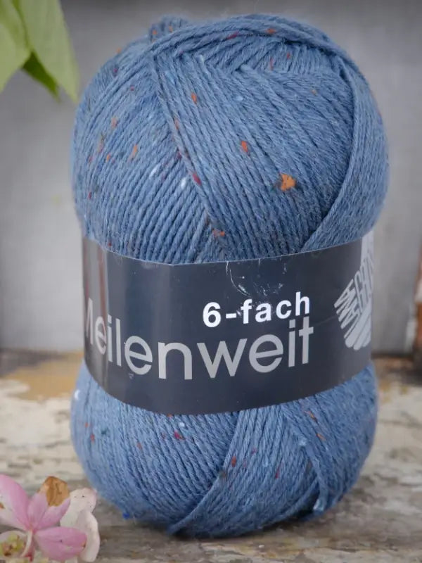 A skein of Lana Grossa Meilenweit 6 Tweed in blue, featuring small colored flecks and labeled as ‘kuusisäikeinen sukkalanka,’ is displayed on a wooden surface alongside a flower and a green leaf.