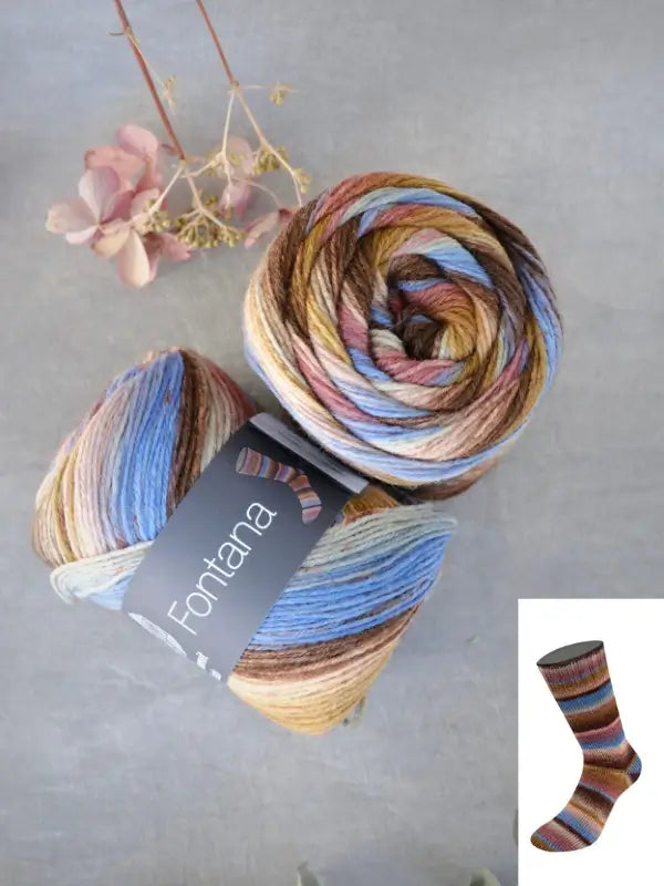 Two skeins of Lana Grossa Meilenweit 100 Fontana, a multicolored yarn in gorgeous shades of brown, blue, and cream, rest on a gray surface next to delicate dried flowers. An inset displays a cozy sock knitted with this yarn, showcasing its vibrant color transitions.