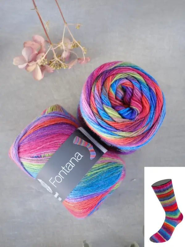 Two skeins of Lana Grossa Meilenweit 100 Fontana rainbow yarn are displayed on a gray surface alongside dried flowers. An inset image features a sock knitted from the same yarn, showcasing its vibrant, multicolored pattern.