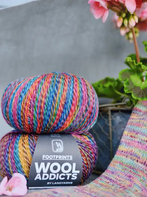 Two skeins of Lang yarns Wool Addicts footprints are stacked beside a potted plant with pink flowers and a piece of knitted fabric made from the same vibrant yarn.