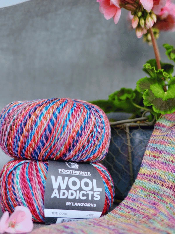 Two skeins of Lang yarns Wool Addicts footprints multicolored sukkalanka yarn are placed beside knitted fabric and a pink-flowered potted plant, with the focus on the yarn and knitting, plants in the background.