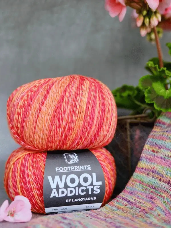 Two balls of Lang yarns Wool Addicts footprints multicolored yarn in red, orange, and yellow are stacked with puuvilla-villasukkalanka and pink flowers nearby on a soft, blurred background.