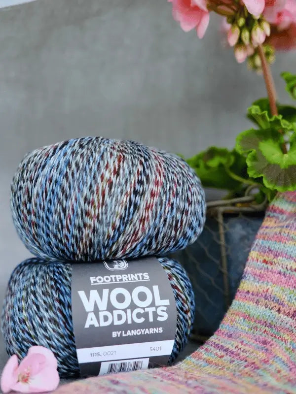 Two skeins of Lang Yarns Wool Addicts Footprints are stacked beside pink flowers and pastel knitted fabric, with a wire basket in the background.