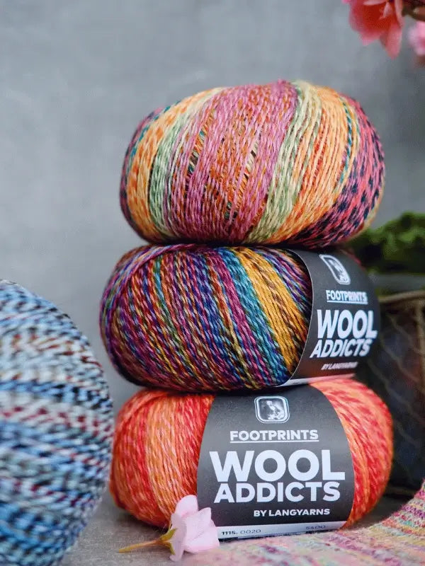 Three colorful balls of Lang yarns Wool Addicts Footprints are stacked, each labeled. In the background, a blurred ball of cotton-wool sock yarn and some plants can be seen.