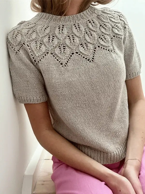 A person wears a light beige Le Knit Sommer Ella neuleohje (ENG), styled with a leaf-patterned shirt in Sandnes Garn Line yarn and pink pants, sitting against a white wall.