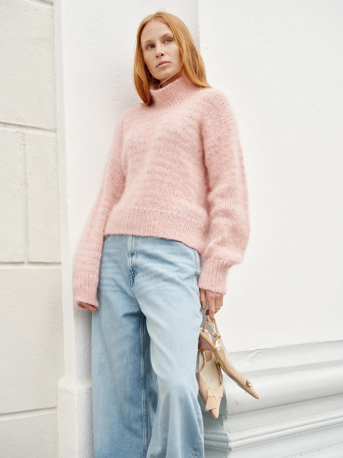A person stands by a white wall, embodying Nordic Chic while holding “Leeni Hoimela – Skandityyli, Ajattomia ohjeita nykyneulojalle” in one hand, dressed in a light pink fuzzy turtleneck sweater and light blue wide-leg jeans.