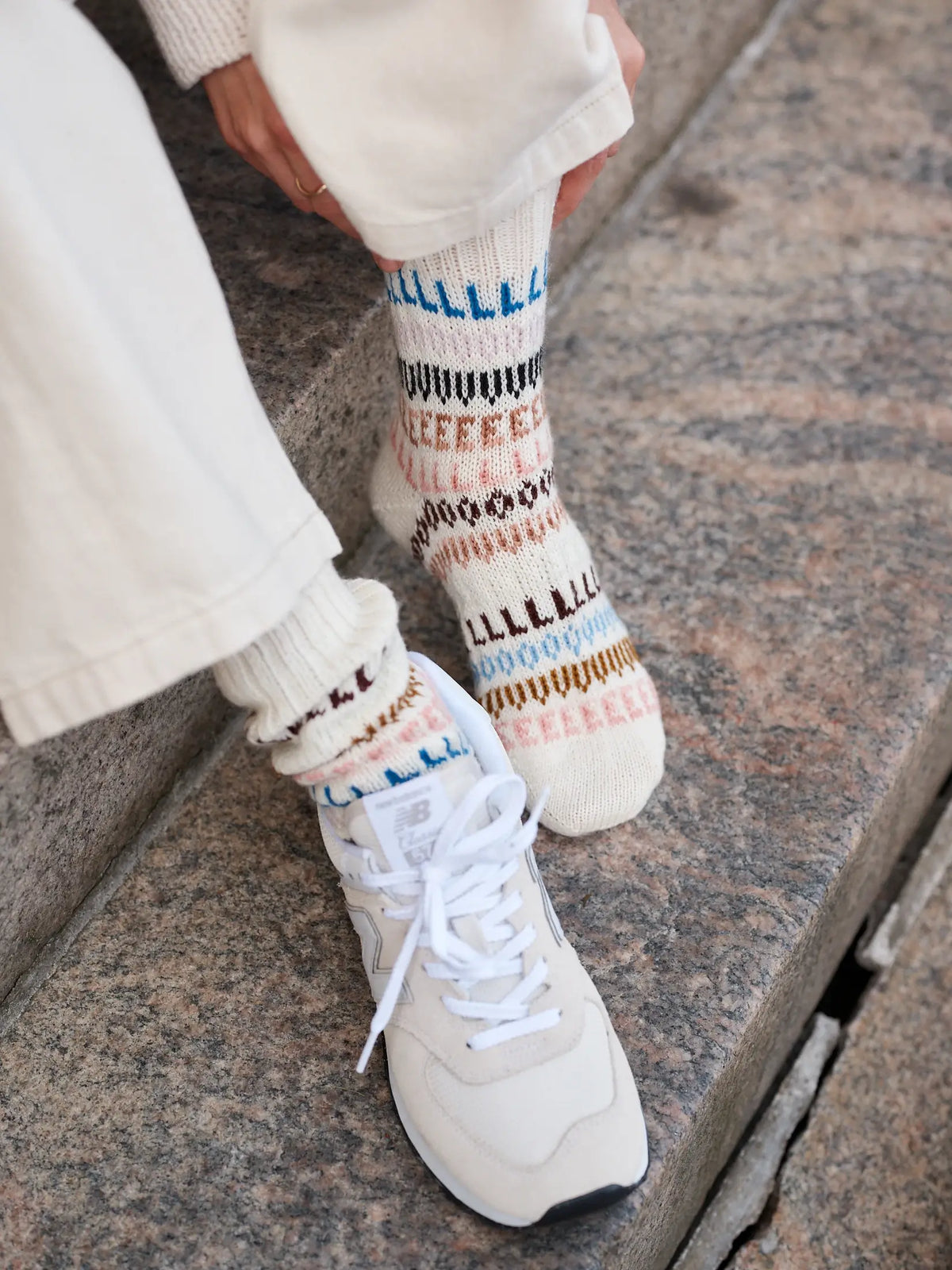 Dressed in cream pants, white sneakers, and vibrant patterned socks featuring repeated L shapes, a person adjusts their sock while sitting on stone steps—a scene that captures the spirit of Leeni Hoimela’s *Skandityyli, Ajattomia ohjeita nykyneulojalle*, effortlessly blending comfort and timeless Nordic chic.