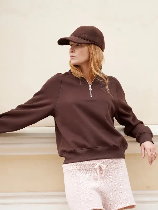A woman in a brown cap and matching zip-up sweatshirt leans against a white ledge, wearing light pink knit shorts with a drawstring—her effortless look perfectly captures the Nordic Chic aesthetic showcased in Leeni Hoimela’s *Skandityyli, Ajattomia ohjeita nykyneulojalle* as she gazes thoughtfully to the side.