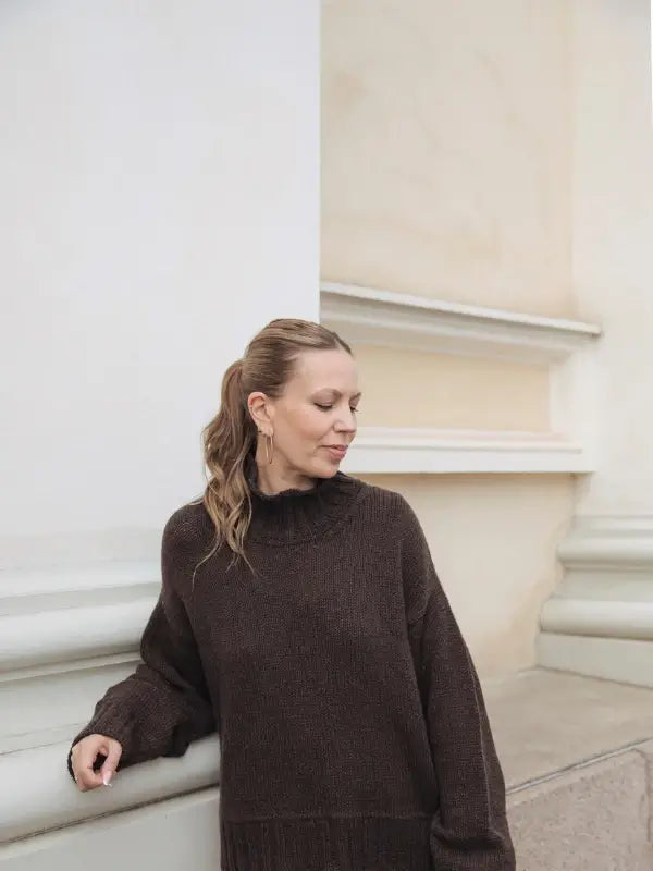 A woman with light brown hair in a ponytail stands near a cream-colored wall, wearing the “Leeni Hoimela – Skandityyli, Ajattomia ohjeita nykyneulojalle” sweater in true Nordic Chic style, gazing down with a relaxed expression.