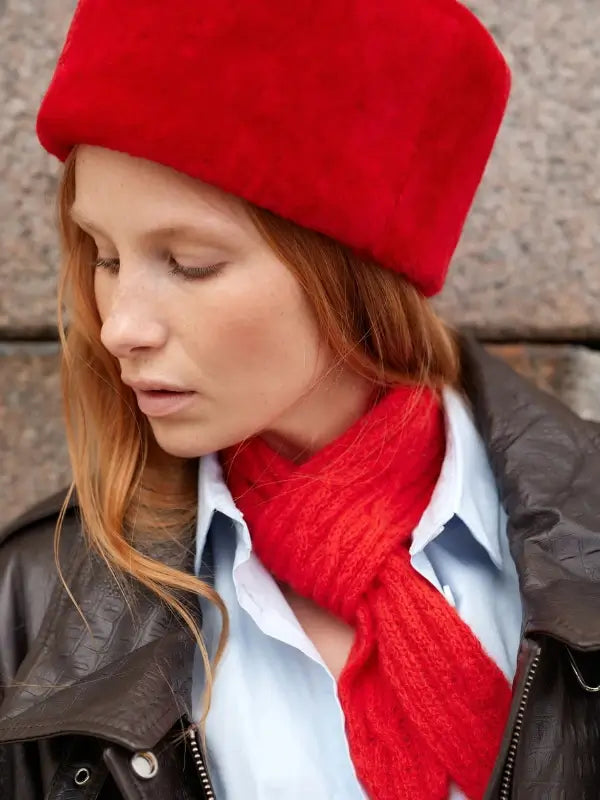 Dressed in Nordic style, a woman with long red hair wears a vibrant red hat and matching scarf as she poses in front of a stone wall, capturing the essence of Leeni Hoimela’s Skandityyli, Ajattomia ohjeita nykyneulojalle. She pairs her light blue shirt and dark leather jacket with timeless Scandinavian accessories while looking down thoughtfully.
