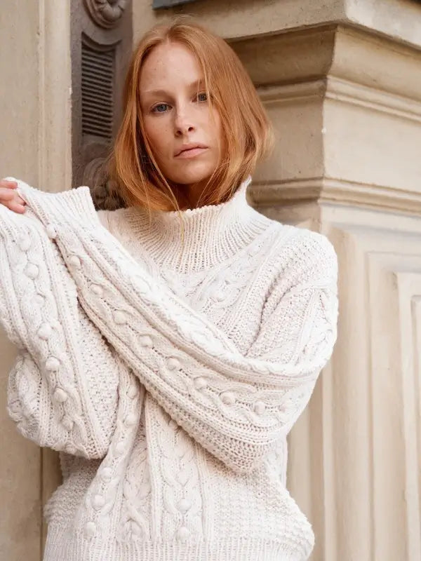 Clutching a copy of Leeni Hoimela – Skandityyli, Ajattomia ohjeita nykyneulojalle, a person with long red hair models effortless Nordic Chic in a chunky, cream-colored cable knit sweater, standing before a beige stone building and gazing neutrally at the camera.