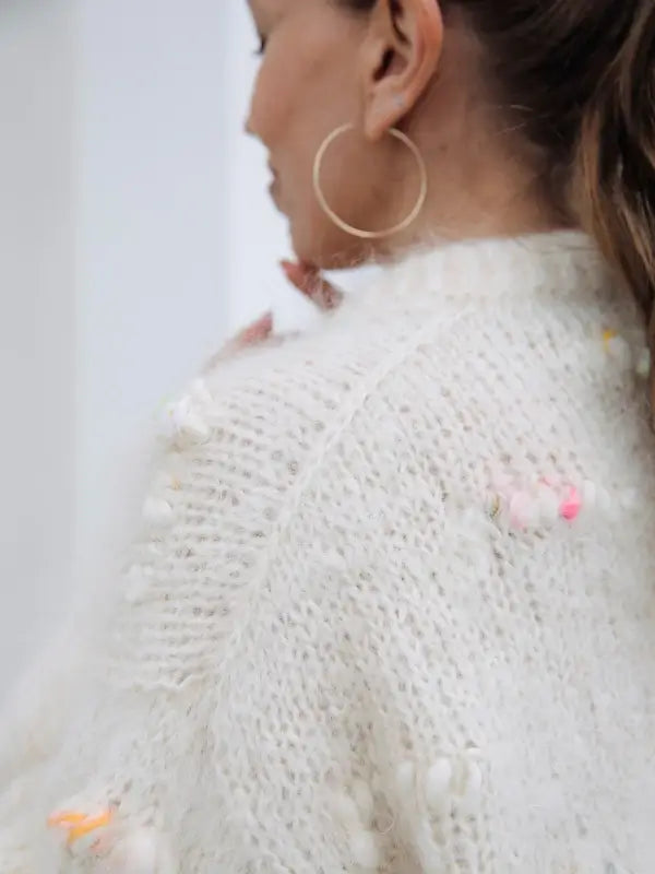 A person with long hair in a ponytail wears a fuzzy white sweater with small colorful accents and large hoop earrings, shown from the side and back—emanating the Nordic Chic style featured in *Leeni Hoimela – Skandityyli, Ajattomia ohjeita nykyneulojalle*.