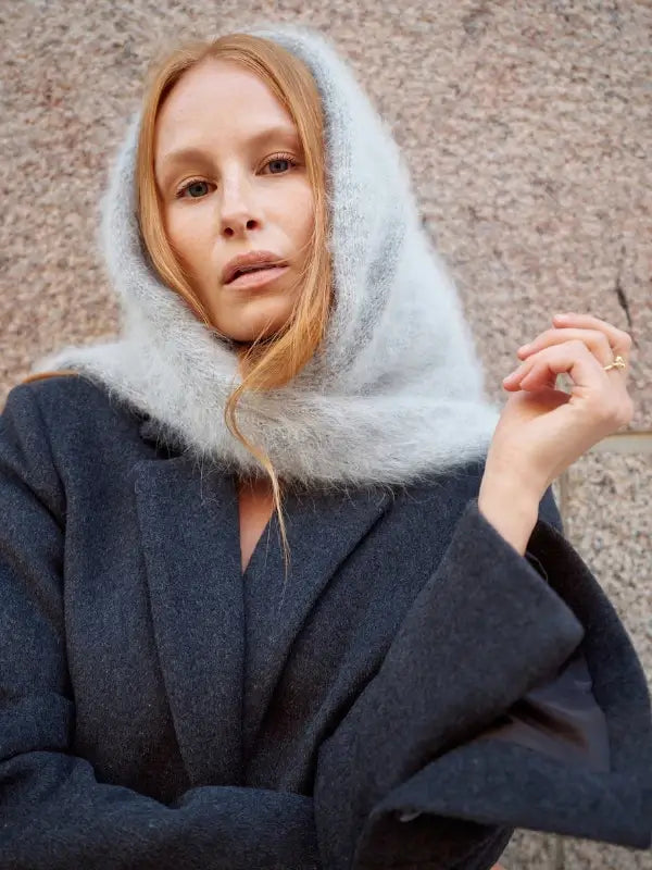 A woman with long red hair, embodying Nordic Chic style, stands in front of a stone wall wearing a fuzzy light gray scarf and a dark coat—the perfect inspiration for the timeless knitting patterns found in *Leeni Hoimela – Skandityyli, Ajattomia ohjeita nykyneulojalle*.