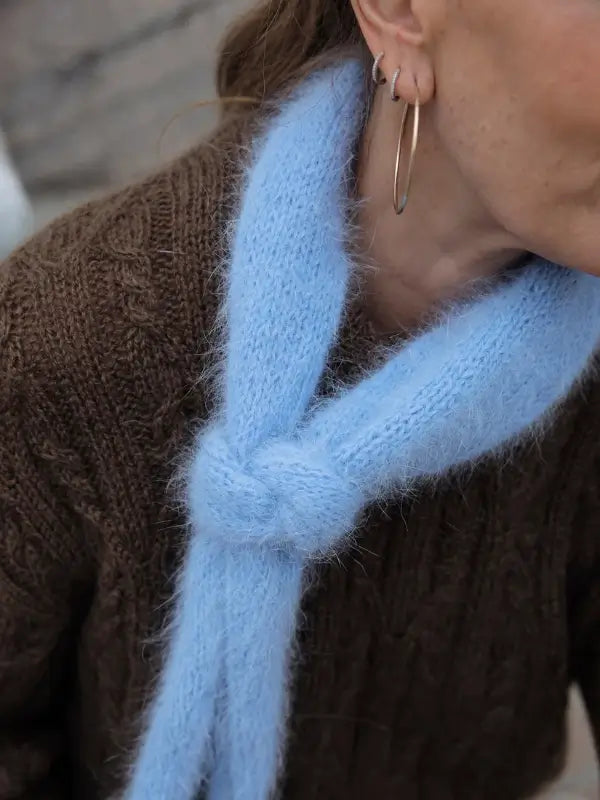 A person wearing a brown knit sweater inspired by “Leeni Hoimela – Skandityyli, Ajattomia ohjeita nykyneulojalle,” perfectly capturing true Nordic Chic style, with a light blue fluffy scarf tied in a knot and large hoop earrings. Only part of the face is visible.