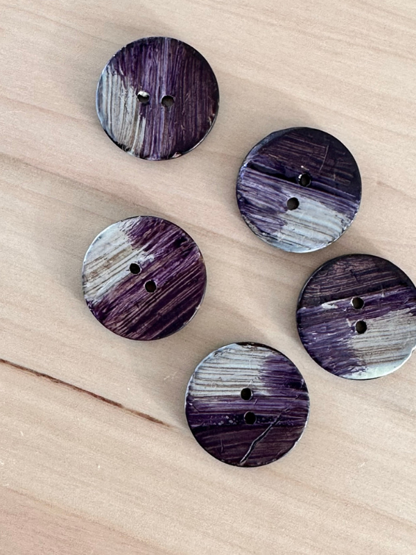 Five round Käsinmaalatut kierrätysnapit 25mm (0454171) with purple and white brushstroke patterns are scattered on a light wood surface, each featuring two holes in the center.