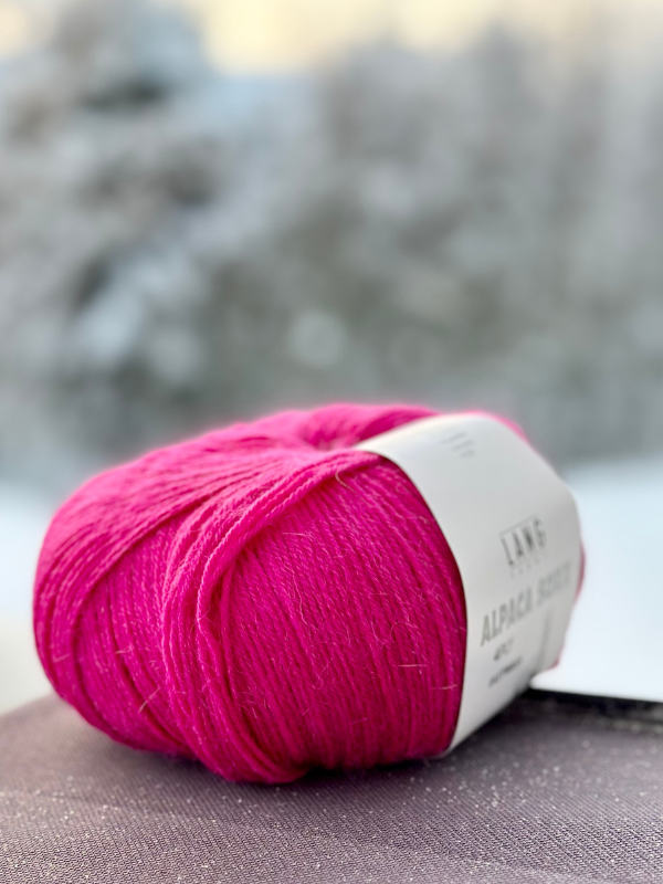 A close-up of vibrant pink Lang Alpaca Soxx 4ply yarn with a white label, paired with the Mittums lapasten ohje (PDF), resting on a dark surface. Blurred snowy trees shine in the background—ideal for your neuleohje project.