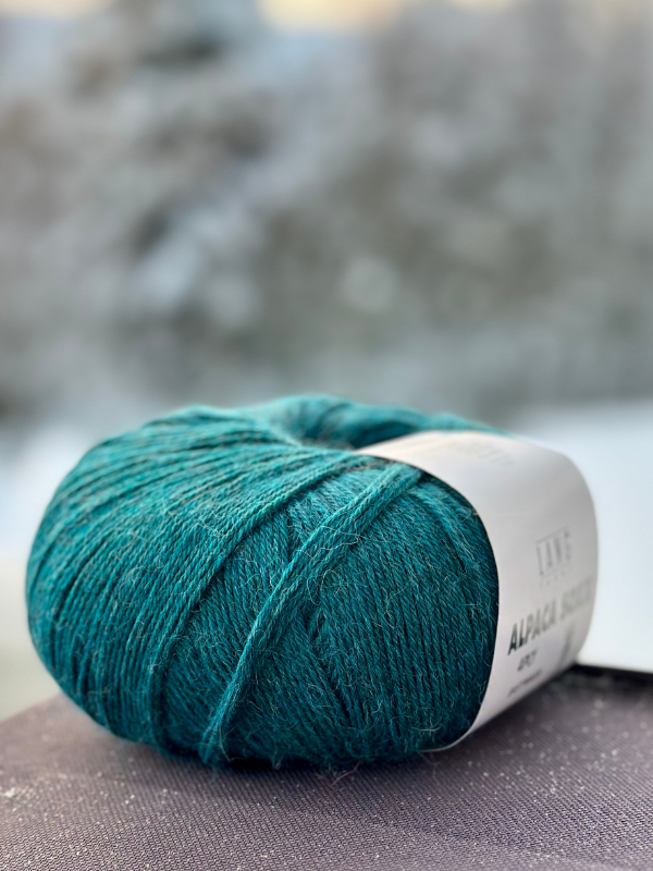 A ball of Lang Alpaca Soxx 4ply yarn for Mittums lapasten ohje (PDF) rests on a surface with its label visible, set against a blurred outdoor background.