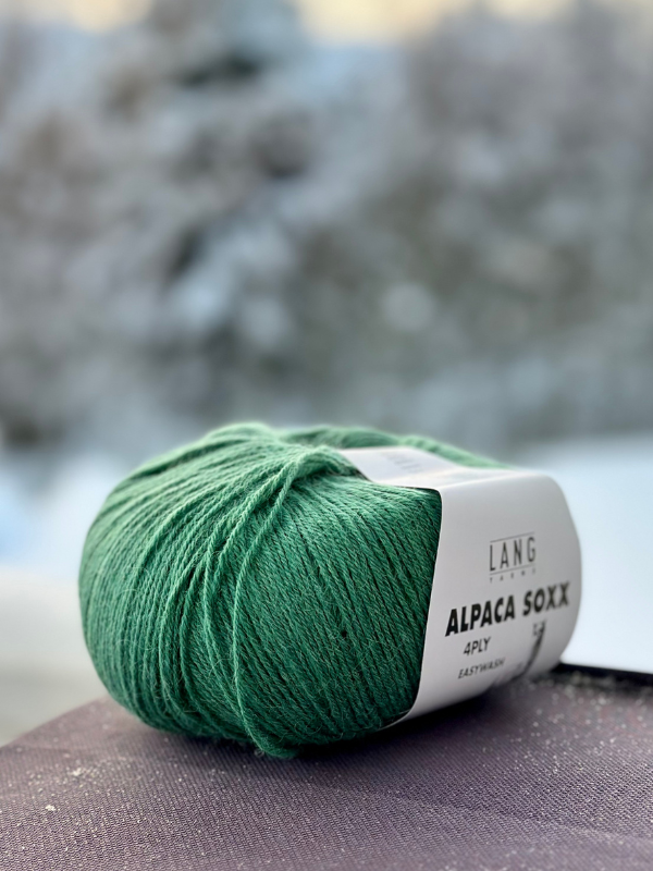 A ball of Lang Alpaca Soxx 4ply sits on a surface with the snowy view behind, paired with Mittums lapasten ohje (PDF)—ideal for your next cozy knitting project or soft mittens.