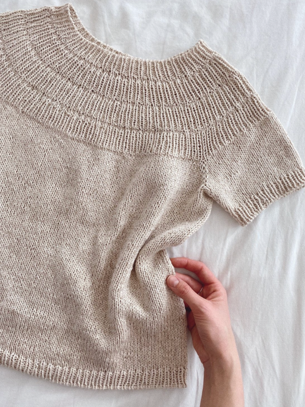 A hand gently holds the hem of a light beige, short-sleeve knitted sweater with a textured yoke—PetiteKnit - Anker’s Summer Shirt neuleohje—laid flat on a white surface.