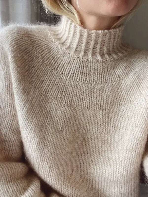 A person is wearing a soft, beige, high-neck knitted sweater. Only the lower half of their face and upper torso are visible. The sweater looks cozy and warm, featuring a ribbed collar and a textured knit pattern—an ideal inspiration for anyone looking for the PetiteKnit - Novice Sweater neuleohje suomenkielinen ohje or searching for beginner-friendly knitting patterns.