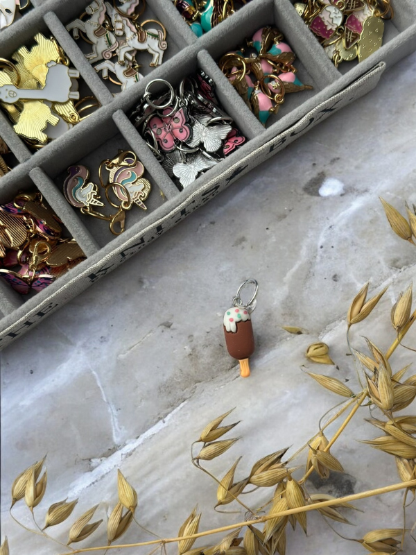 A marble surface holds a chocolate popsicle-shaped keychain with white drizzle, beside a beige tray filled with KNITS by cindy ekman silmukkamerkit for neulonta, and dried oat stalks in front.