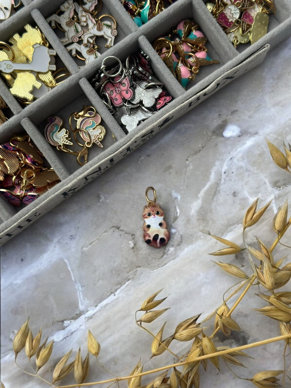 A box of KNITS by cindy ekman silmukkamerkit sits on a marble surface. A cute cat paw charm lies beside it—perfect for neulonta. Dried oat stalks decorate the bottom right corner.