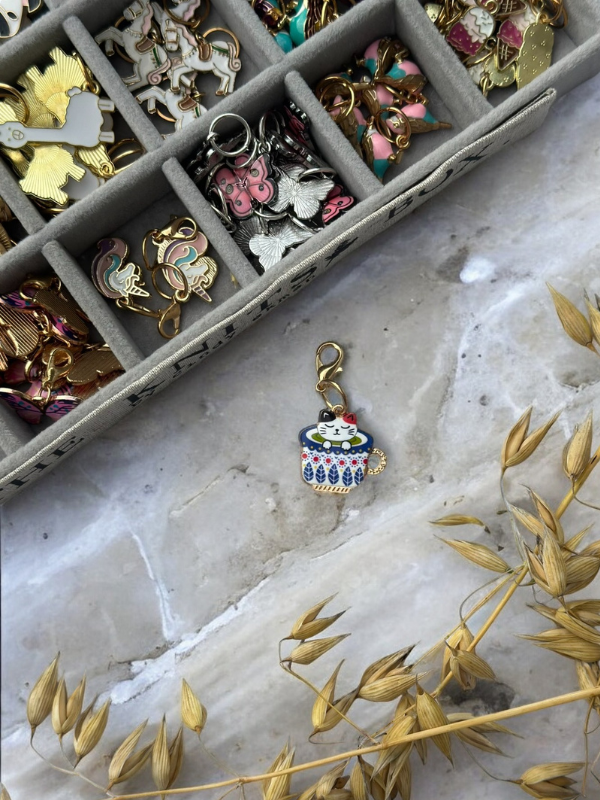 A colorful enamel cat-in-a-teacup charm rests on marble beside a beige box filled with KNITS by cindy ekman silmukkamerkit, metal charms, and dried wheat stems.