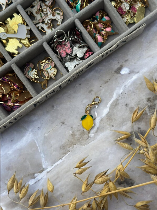 A tray of colorful KNITS by cindy ekman silmukkamerkit, enamel charms, and keychains is displayed on marble; a yellow lemon-shaped charm with a green leaf rests nearby beside dried golden grass.