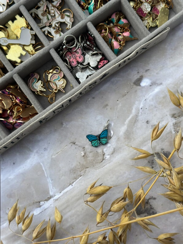 A turquoise butterfly charm rests on marble beside a box of KNITS by cindy ekman silmukkamerkit, colorful enamel pins, and dried golden grass.