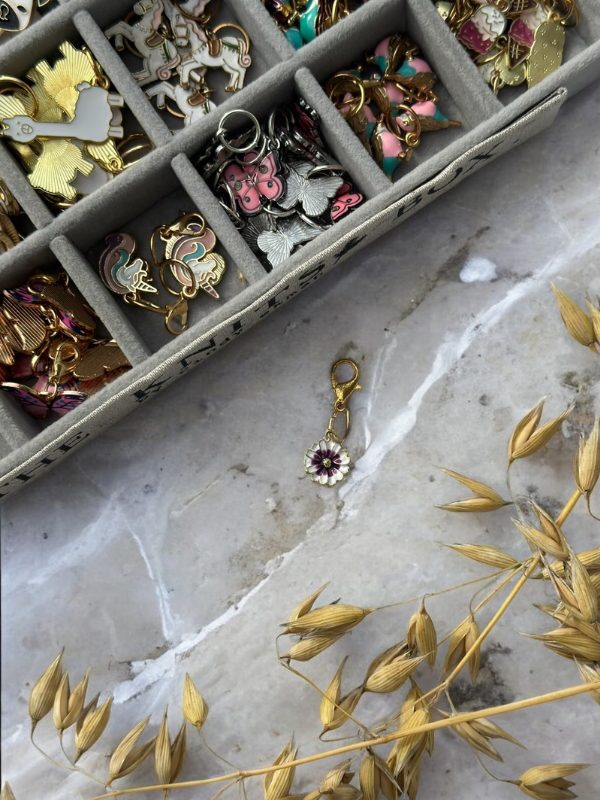 The KNITS by cindy ekman silmukkamerkit jewelry organizer, filled with colorful charms, rests on marble. A separate flower-shaped charm with a clasp lies nearby, accented by dried golden grasses in the corner.