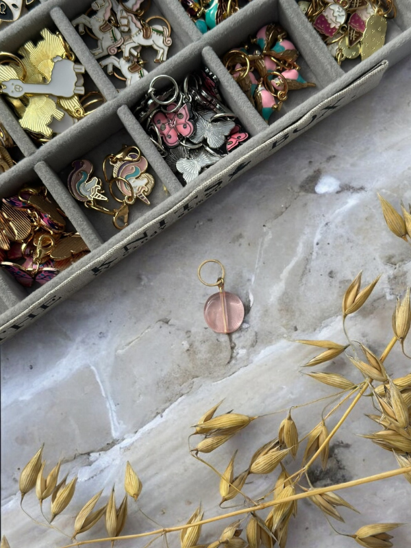 A pink circular pendant with a gold loop rests on marble near dried wheat stalks; above it is a tray of colorful and gold jewelry charms, including KNITS by cindy ekman silmukkamerkit—perfect for neulonta projects.