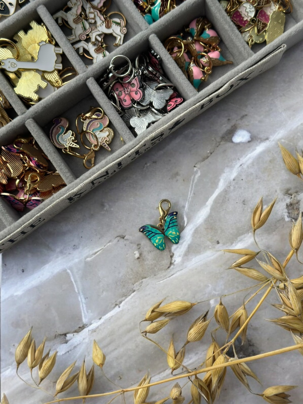 A turquoise butterfly charm rests on marble next to a beige box containing KNITS by cindy ekman silmukkamerkit for neulonta, with dried golden grass in the corner.