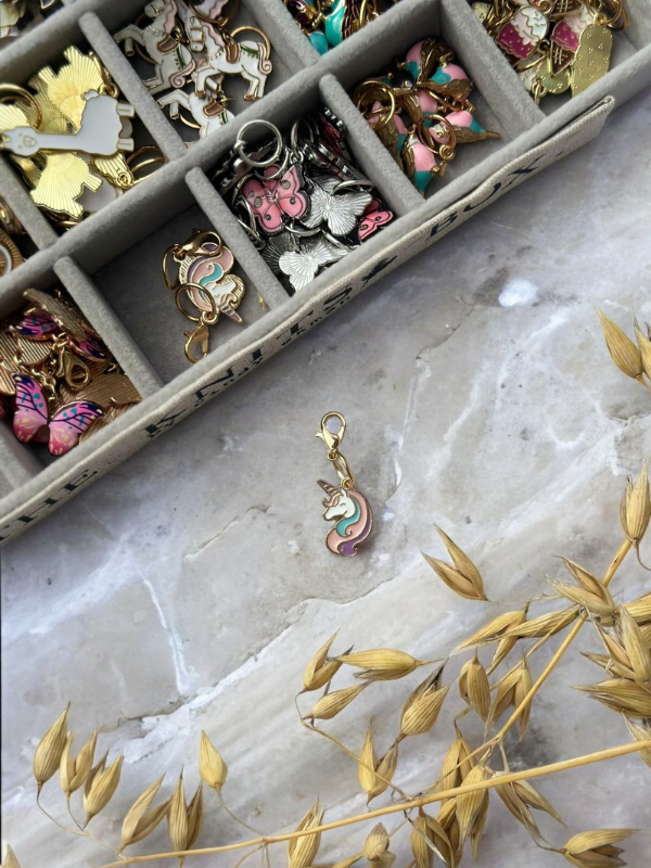 KNITS by cindy ekman silmukkamerkit enamel charms, including a pastel cat-shaped one, are neatly arranged in a gray tray on marble. Dried oat stalks are visible at the bottom right.