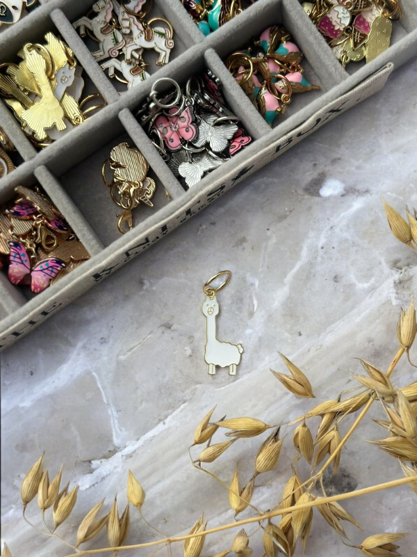A gold llama-shaped KNITS by cindy ekman silmukkamerkit keychain rests on a marble surface near dried plants, with a tray of colorful neulonta pins and other animal-shaped stitch markers in the background.