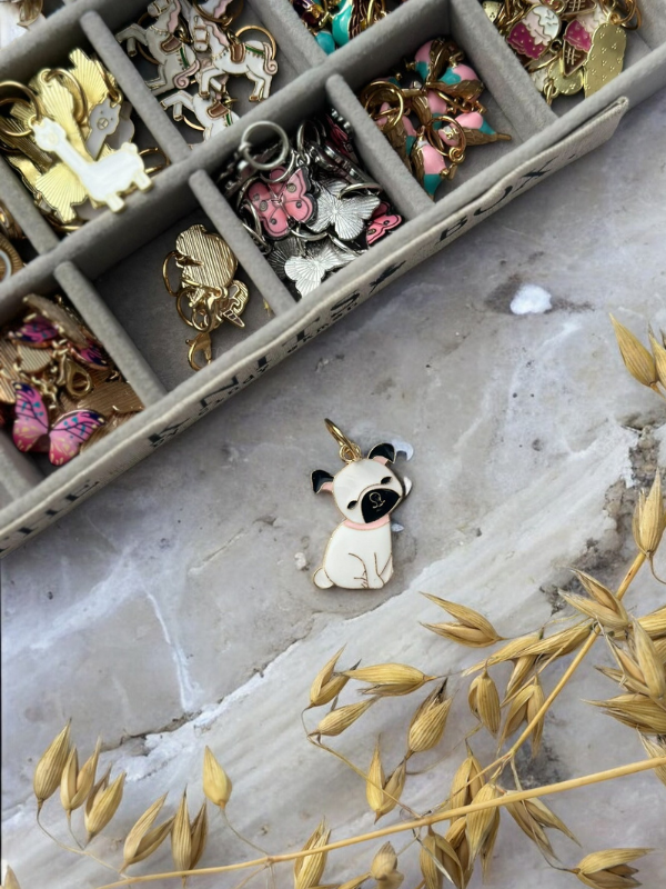 Flat lay of a beige and black pug-shaped enamel charm on marble, with a box of KNITS by cindy ekman silmukkamerkit and dried yellow grass in the corner.