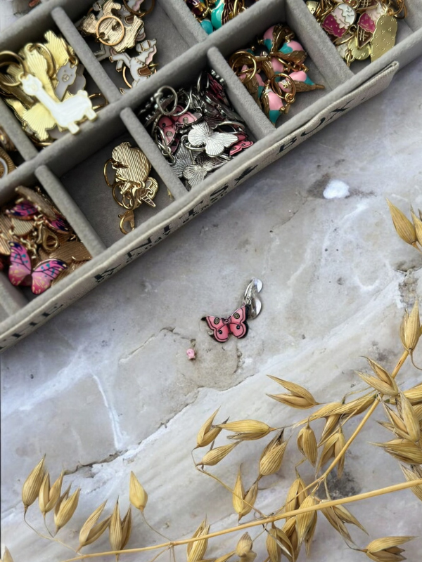 A KNITS by cindy ekman silmukkamerkit jewelry box with colorful butterfly and assorted charm markers rests on marble. One pink butterfly marker and a bead lie beside it near dried golden grass—perfect for neulonta projects.