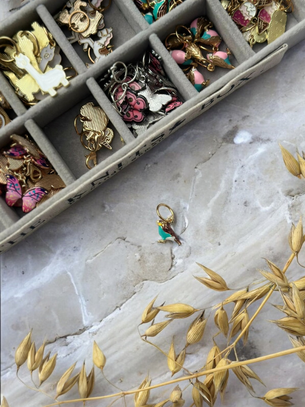 The KNITS by cindy ekman silmukkamerkit jewelry organizer, decorated with colorful enamel charms, rests on marble. A gold charm with green and white accents lies outside the organizer, with dried golden grass in the lower right corner.
