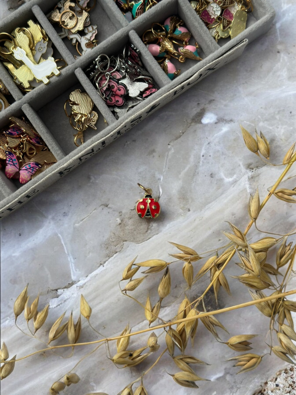 A gold tray displays KNITS by cindy ekman silmukkamerkit with colorful pins on a gray marble surface. A red ladybug charm rests beside the tray, near dried golden grass.